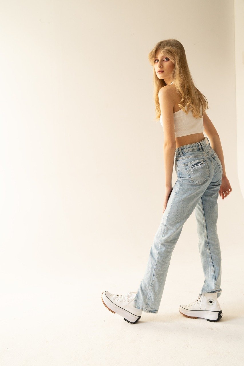 young woman, sporty outfit, denim style, white top, casual sneakers, blond hair, casual style, youthful look, fashion model, long hair, elegant appearance, fashion photoshoot, model standing, white room, youth fashion, natural light, wavy hair, young girl, floor pose, long legs, light blue jeans