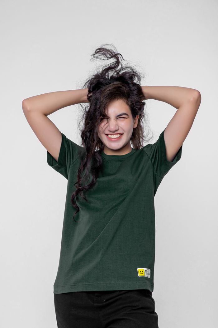 Cheerful young woman in green t-shirt smiling with hands in hair against white background.