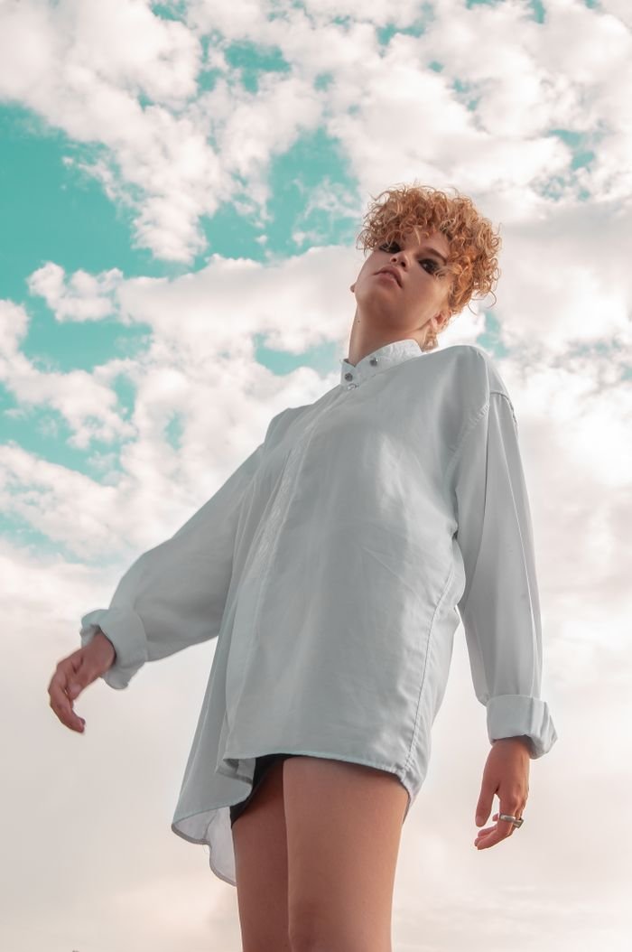 Low angle of serious female with makeup and curly hair standing against cloudy sky and looking away